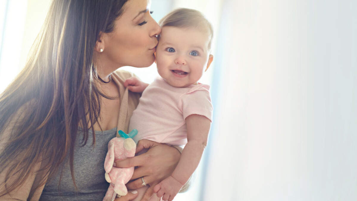 Mother, bonding and forehead kiss for baby girl on in house living room, nursery or family home bedroom in trust or love. Smile, happy and infant child with mom in support, security or safety comfort.