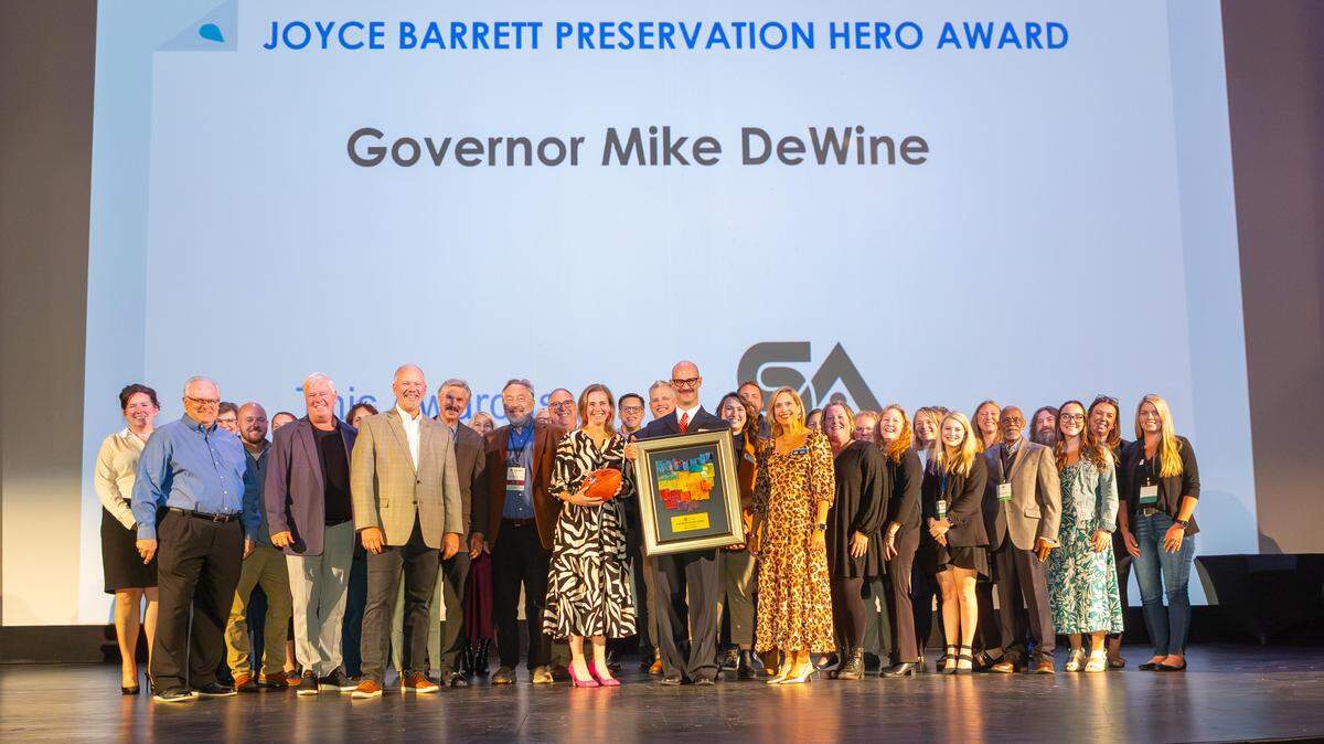 Matt Wiederhold, Executive Director of Heritage Ohio, presents the Joyce Barrett Historic Preservation Hero award to Ohio History Connection President Megan Wood, on behalf of Governor Mike DeWine. They are joined on stage by the Heritage Ohio Board and Ohio Main Street Programs Executive Directors. The award is a custom work of art by Heritage Ohio Board Chair and artist, Dave Williams, of Dayton.