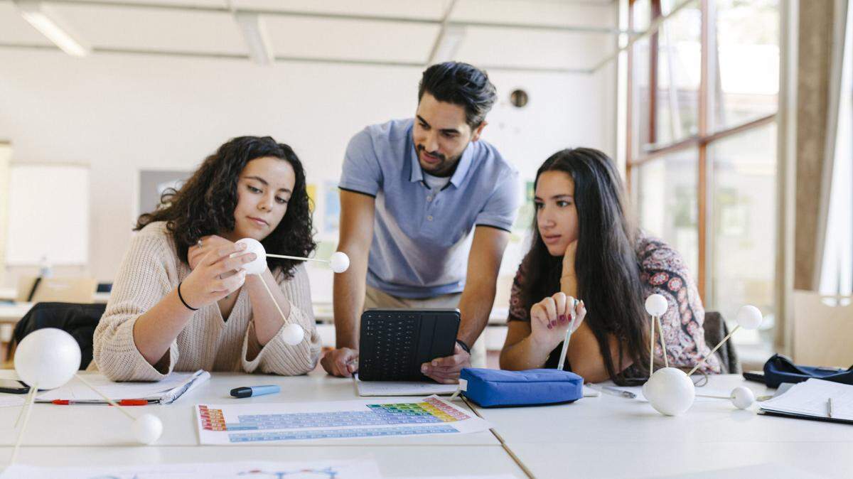 A high school science teacher holding a digital tablet and explaining a chemistry model to two of his students during class.