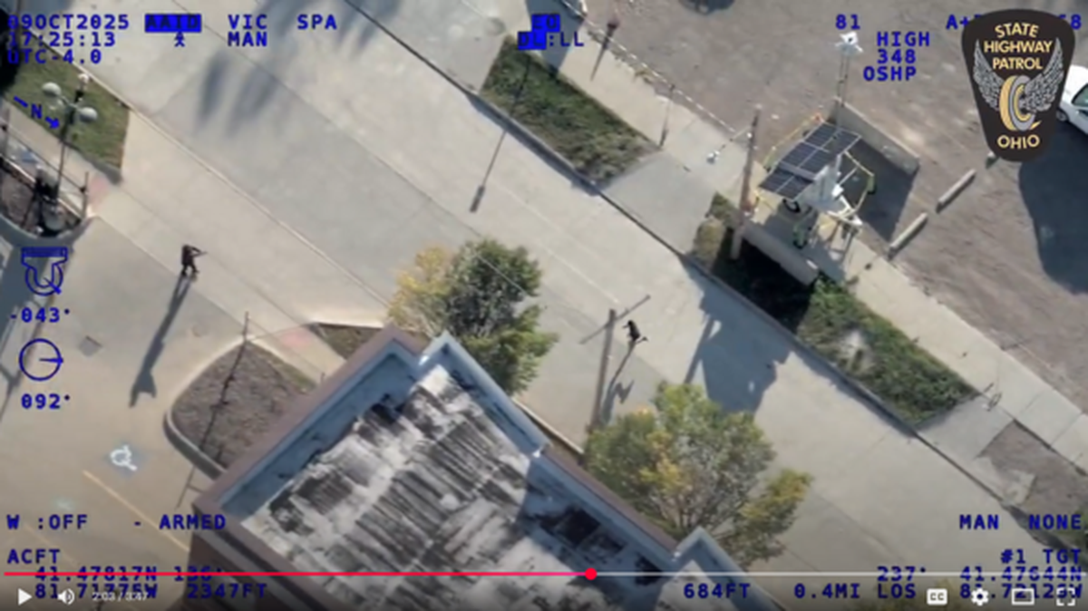 Aviation Video Still: Law enforcement officer stands to right of frame with suspect running in middle of road.