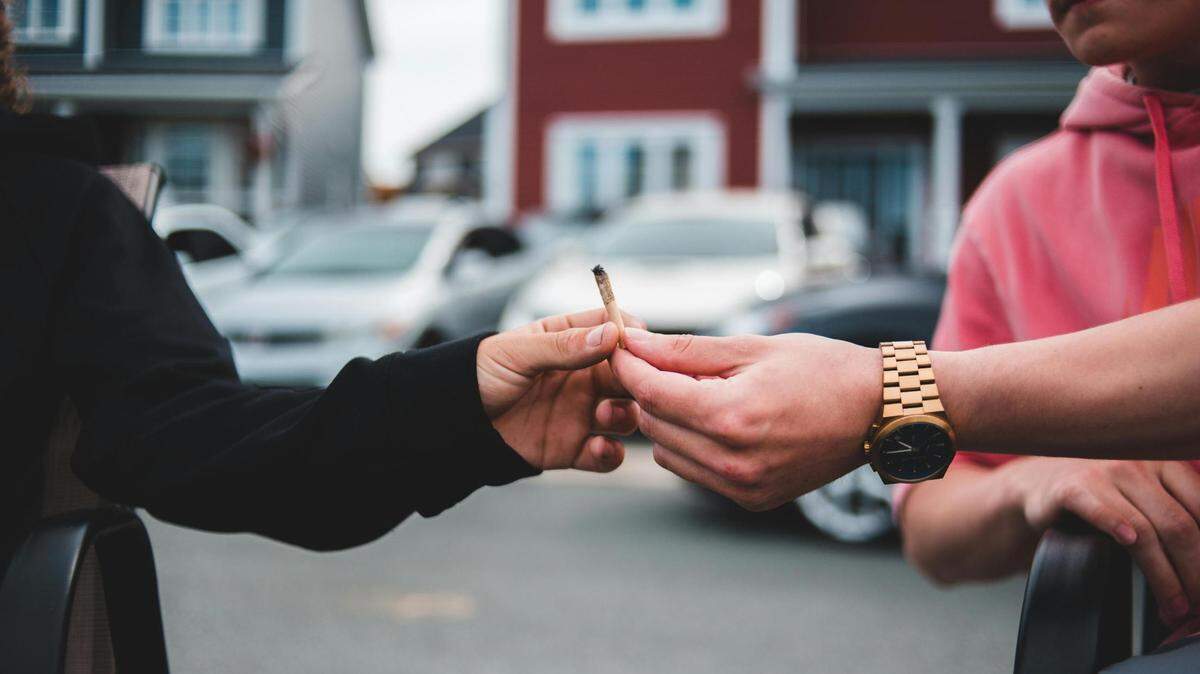 A man is passing a joint of marijuana (cannabis, pot) to another person with cars and homes in background.